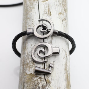 May include: A black braided leather bracelet with a silver-colored spiral clasp. The clasp is open, showing the separate pieces. The bracelet is displayed on a white and brown wooden surface.