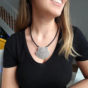 Op de afbeelding: Een zilverkleurige schelphanger ketting met een zwart koord. De hanger, met geribbelde details, is bevestigd aan een zwart koord. De ketting wordt gedragen over een zwarte V-hals top.