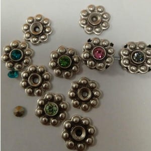 Puede incluir: Surtido de adornos metálicos en forma de flor de color plateado con piedras de cristal de varios colores en el centro. Las piedras de cristal son verdes, rosas, azules y transparentes.