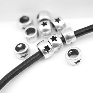 May include: Silver-colored metal beads with star cutouts are strung on a black leather cord. Several loose beads are scattered around the cord. The beads have a worn, vintage appearance, and are set against a white background.