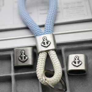 May include: A white and blue rope keychain with three silver metal anchor charms. The charms are square shaped and have a raised anchor design.