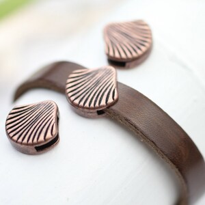 Puede incluir: Primer plano de una pulsera de cuero marrón con tres cierres en forma de concha de color cobre. Los cierres tienen un diseño detallado y acanalado. La pulsera está sobre una superficie blanca, con los cierres enfocados.