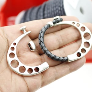 Puede incluir: Un brazalete de plata con una correa de cuero trenzada y un diseño de cierre único. El cierre tiene forma de herradura con varios agujeros. El brazalete se muestra desmontado con el cierre y la correa separados.