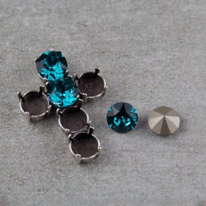 May include: A silver-toned cross pendant with teal-colored gemstones. Two loose gemstones are also visible, one teal and one metallic gold. The cross is set against a gray background.