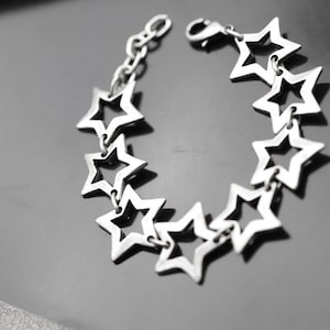 Op de afbeelding: Zilverkleurige armband met een reeks open sterren die met elkaar verbonden zijn. De armband heeft een karabijnsluiting en een kleine ketting om de lengte aan te passen. De sterren zijn gelijkmatig verdeeld.
