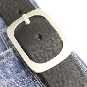 Puede incluir: Un cinturón de cuero negro con una hebilla ovalada plateada. El cinturón está abrochado alrededor de un par de pantalones vaqueros azules.