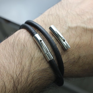 May include: A black leather bracelet with a silver magnetic clasp. The bracelet is double-wrapped around a wrist.