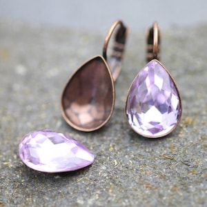 Puede incluir: Un par de pendientes colgantes de cobre con un cristal de color púrpura claro en forma de lágrima. Los pendientes se muestran sobre una superficie texturizada gris oscuro.