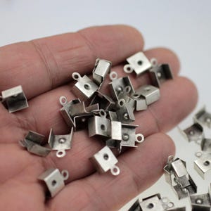 May include: A pile of silver-toned metal square-shaped clasps with a loop on each one. The clasps are designed to be used for jewelry making.
