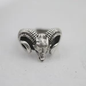 May include: A silver-coloured metal ring featuring a detailed ram skull design. The centrepiece displays the head of a ram with prominent, textured curved horns extending to the sides. The metal has a darkened, oxidised finish in the recessed areas.