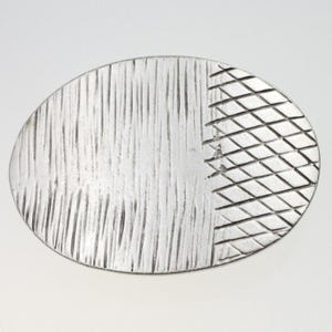 Op de afbeelding: Een zilveren ovaalvormige broche met een textuurontwerp. De ene helft van de broche heeft een verticaal lijnenpatroon, terwijl de andere helft een kruispatroon heeft.