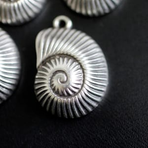 Peut inclure: Pendentif en métal argenté en forme de coquille avec un motif en spirale. Le pendentif a une surface texturée détaillée.