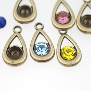 Könnte beinhalten: Nahaufnahme von mehreren tropfenförmigen Schmuckteilen in antik-bronzenem Finish. Einige Teile weisen farbige, runde Edelsteine in Rosa, Blau und Gelb auf. Die Teile haben oben eine Schlaufe zum Befestigen an anderen Schmuckkomponenten.