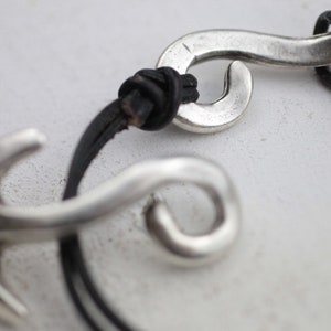 Puede incluir: Un brazalete de plata con un cordón de cuero negro. El encanto es un diseño de remolino estilizado.