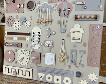 May include: A wooden activity board with various textures and activities for children. It features a clock, a zipper, a shoe with laces, a hot air balloon, a cloud with beads, a light switch, a keyhole, a chain, a gear, a maze, and a door with a latch. The board is painted in shades of gray and pink and has the name "LENA" written on it.