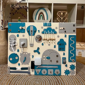 Peut inclure: Un tableau d'activités en bois pour tout-petits avec une variété de textures et d'activités, notamment une fermeture éclair, un loquet, une horloge, une chaussure avec des lacets, une montgolfière, un nuage avec des perles, une porte avec un loquet, un labyrinthe, un engrenage et un trieur de formes. Le tableau est peint dans des tons de bleu, de gris et de blanc et porte le nom "IRIS".