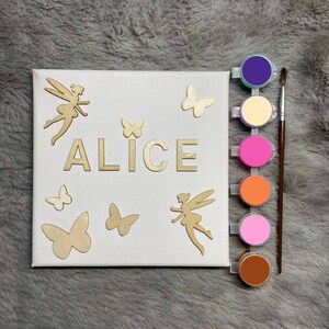 Op de afbeelding: Een wit canvas met de naam "ALICE" in gouden letters, omringd door gouden fee- en vlinderuitsnijdingen. Het canvas wordt vergezeld door zes verfpotjes in verschillende kleuren: paars, crème, roze, oranje, lichtroze en bruin.