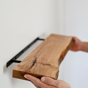 Op de afbeelding: Een persoon installeert een houten plank met zwarte metalen beugels. De plank is gemaakt van natuurlijk hout met een levende rand en wordt vastgehouden door de handen van de persoon.