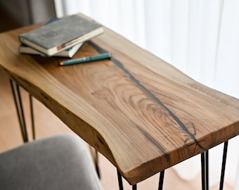 May include: A wooden table with a live edge top and black hairpin legs. The table has a natural wood finish and a black epoxy resin inlay. There are three books and a pen on the table.