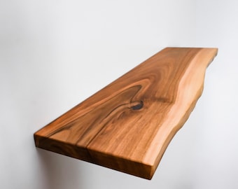 May include: A floating wooden shelf with a natural edge. The shelf is made of a warm-toned wood with visible grain and a knot. The shelf is mounted on a white wall, showcasing its unique shape and texture.
