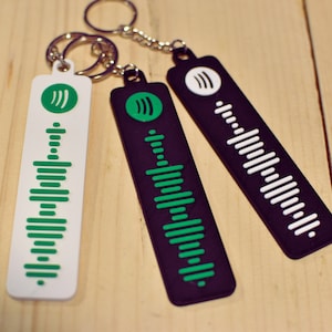 Op de afbeelding: Drie sleutelhangers met een geluidgolfontwerp. Eén sleutelhanger is wit met groene geluidgolven, één is zwart met groene geluidgolven en één is zwart met witte geluidgolven.