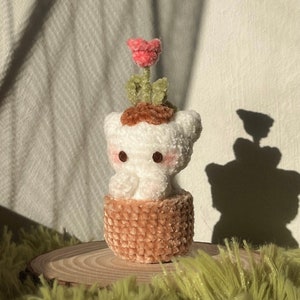 May include: A crocheted plush toy shaped like a potted plant with a white body, brown pot, and a pink flower on top.