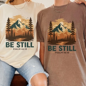 Könnte beinhalten: Zwei T-Shirts, eines in Creme und eines in Braun, mit einem Berg- und Waldmotiv. Der Text "BE STILL" und "PSALM 46:10" sind darunter gedruckt. Die Shirts werden von zwei Personen getragen.