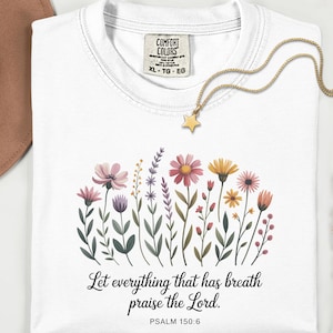 以下が含まれることがあります： 白いComfort ColorsのTシャツ。花柄のデザインと「Let everything that has breath praise the Lord. Psalm 150:6」のテキスト。金の星のペンダントネックレスがシャツの上にあります。茶色の帽子が左上にあります。