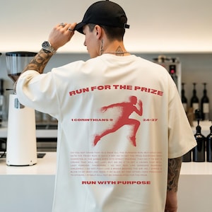 May include: Off-white t-shirt featuring the red text "RUN FOR THE PRIZE" and a red runner graphic. The shirt also displays the text "1 CORINTHIANS 9 24-27". Further text is printed below the graphic, in a bold red font.