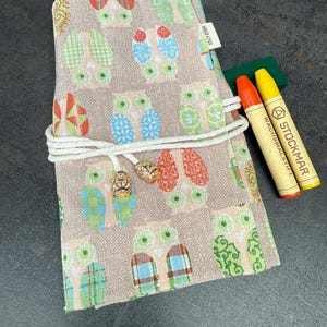 May include: A rolled-up, owl-patterned pencil case with a white cord closure and gold-colored charms. Two crayons, one orange and one yellow, are next to the case. The pencil case has a beige background with colorful owl designs.