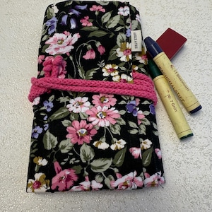 May include: A floral fabric pencil case with a pink knitted band. The case is black with pink, white, and purple flowers. Two crayons and a small red eraser are next to the case. The text "SILLY STUFF" is visible on the case.