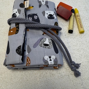 May include: A gray fabric roll-up pouch with a dog print design. The pouch is tied with a gray rope and has a label that says "Silly Stuff". The pouch is sitting on a white surface with two yellow and brown crayons and a red rectangular object.