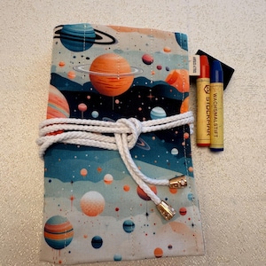 May include: A fabric pencil case with a colorful space-themed design featuring planets and stars. The case is tied with a white rope and gold-colored accents. Two crayons with the text "WACHSMALSTIFT" are visible.