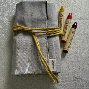 May include: A beige fabric crayon roll with a yellow cord closure, holding three Stockmar crayons in yellow, red, and burgundy. The roll is decorated with gold polka dots and the label reads "SILLY STUFF."