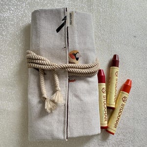 May include: A beige canvas pencil case tied with a rope, featuring a small bird illustration and the text "SILLY STUFF." Three red and orange STOCKMAR crayons are next to the case.