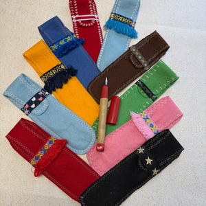 Peut inclure: Une collection de dix étuis à stylos en feutrine de différentes couleurs, dont rouge, bleu, jaune, vert, rose, marron et noir, disposés en éventail. Chaque étui présente des coutures décoratives, des franges ou des bandes de tissu à motifs. Un stylo rouge et bois est placé au centre.