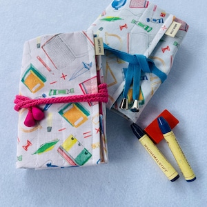 May include: Two white fabric pencil cases with a school supplies print. The cases are tied with blue and pink ribbon. Two blue and yellow crayons with the text "WACHSMALSTIFT" are on a light blue surface.