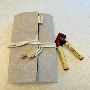 May include: A beige fabric crayon case with gold star accents, tied with a white cord and gold-colored clasps. The label reads "SILLY STUFF". Two red and brown crayons are next to the case, one labeled "STOCKMAR" and the other "WACHSMALSTIFT".