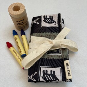 May include: A black and white fabric roll-up pouch with a white ribbon tied around it. The pouch has a label that says "Silly Stuff" and features a pattern of elephants. The pouch contains a set of crayons and a wooden pencil sharpener.