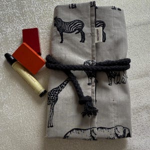May include: A gray fabric pencil case with black animal illustrations, including zebras and giraffes, tied with a black rope. Includes red and orange wooden blocks and a black and yellow pencil.