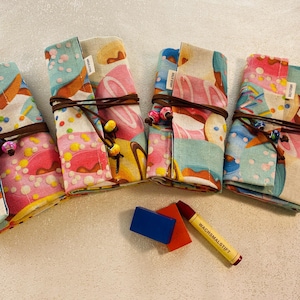 May include: Four colorful fabric pencil cases with a donut and sprinkles pattern. Each case has a brown leather strap and a small tag with the word "Made with Love" printed on it. The cases are arranged in a row on a white surface. There is a blue eraser and a yellow crayon with the text "Wachsmalstift" on it.
