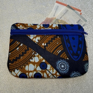 Peut inclure: Un petit porte-monnaie rectangulaire avec une fermeture éclair bleue et un imprimé africain vibrant dans les tons marron, bleu et blanc. Le porte-monnaie est ouvert, révélant plusieurs billets en euros. Conçu pour contenir de la monnaie et de petits objets.