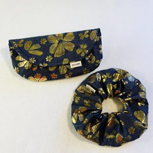 Op de afbeelding: Een donkerblauwe stoffen clutch met een gouden bloemenpatroon en de tekst "Silky Stoff" aan de voorkant. Een bijpassende donkerblauwe stoffen scrunchie met een gouden bloemenpatroon.