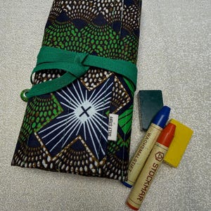 May include: A rolled-up fabric pencil case with a green and blue geometric pattern, tied with a green ribbon. The case is next to two crayons and a small yellow eraser. The text "SILLY STUFF" is visible on the case.
