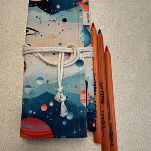 May include: A rolled-up pencil case with a space-themed pattern, featuring planets and stars in shades of blue, orange, and white. The case is tied with a white rope and holds several colored pencils. The text "SILLY STUFF" is visible on the case.