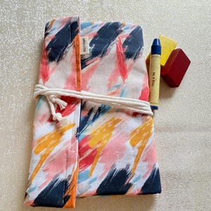 May include: A colorful fabric roll-up case with a brushstroke design in pink, orange, blue, and black, tied with a white rope. The case holds a blue crayon and two other crayons. The text "SILLY STUFF" is visible on the case.