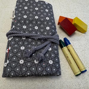 May include: A gray and white patterned fabric pencil roll with a rope closure. The roll is filled with crayons and is sitting on a white surface. The text "Silly Stuff" is printed on the roll.