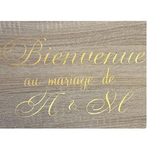 Peut inclure: Un panneau rectangulaire avec une finition en grain de bois brun clair portant le texte doré en cursive Bienvenue au mariage de A & M. Le lettrage est centré sur la surface, affichant un style de police script décoratif sur trois lignes.