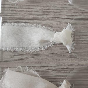 May include: Three clear acrylic circular tags, each featuring a piece of white fabric ribbon tied through a small rectangular hole. The ribbons have frayed, raw edges and are knotted securely to the smooth, transparent acrylic discs.