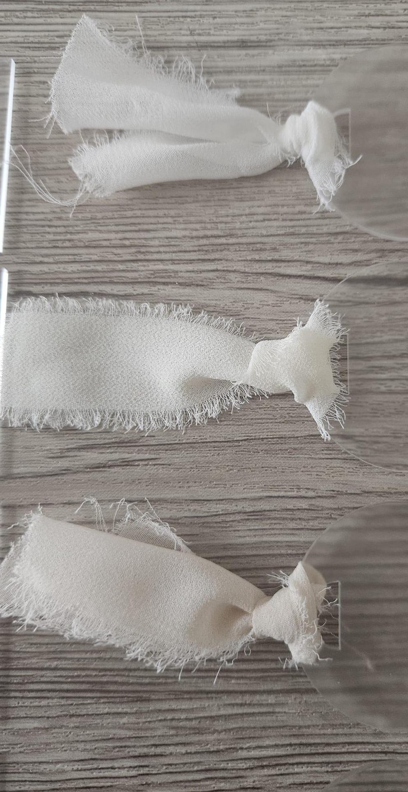 May include: Three clear acrylic circular tags, each featuring a piece of white fabric ribbon tied through a small rectangular hole. The ribbons have frayed, raw edges and are knotted securely to the smooth, transparent acrylic discs.